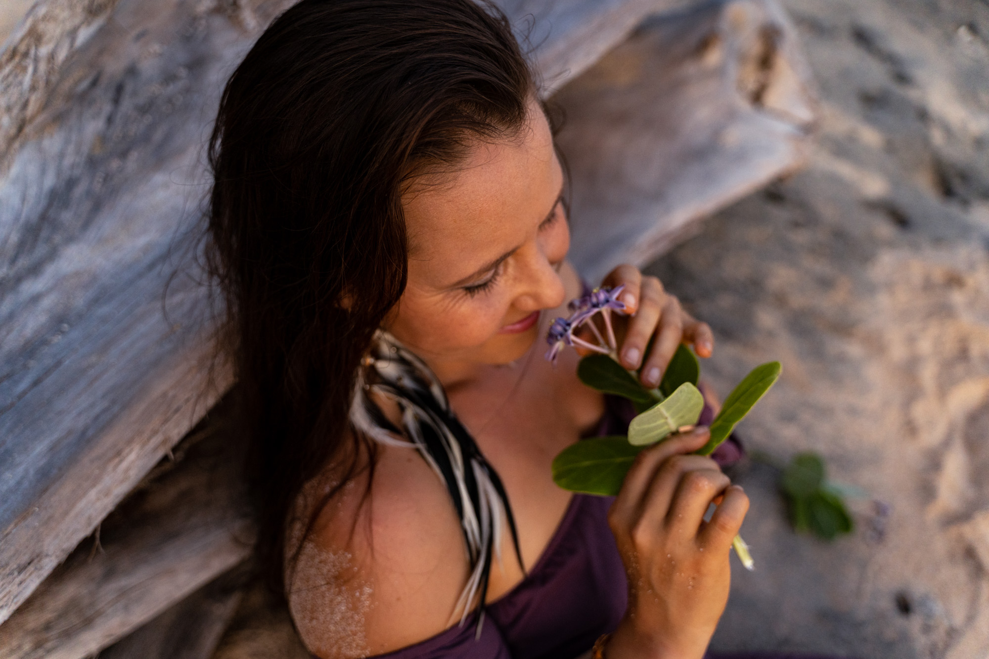 Sara Adolfsen somatic educator for women is dressed in purple, sitting on a beach, back against an old log, smiling and smelling a purple flower